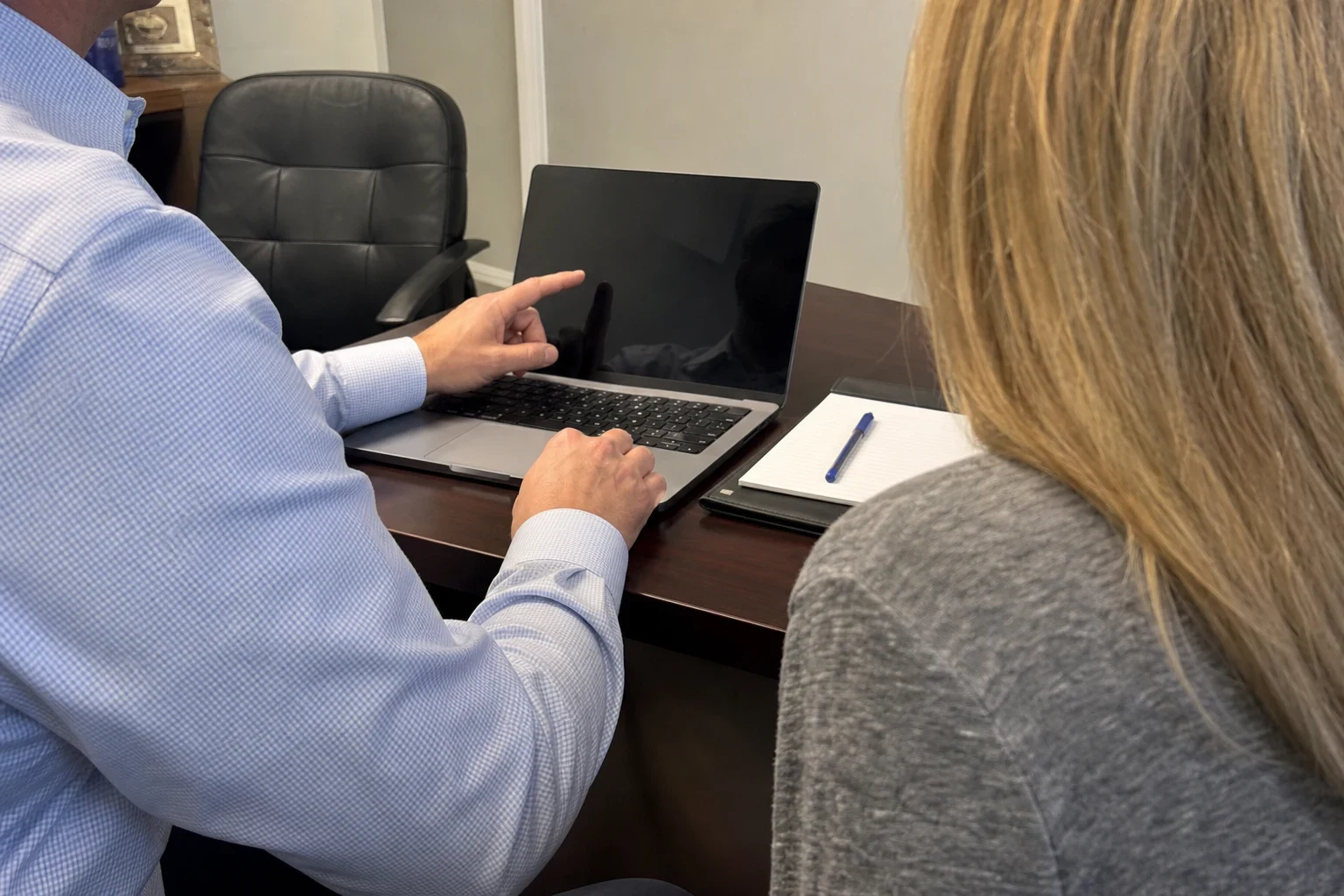 Agent in a blue checked shirt points at a laptop on a wood desk while a client with long blonde hair looks on, a notepad and pen beside them