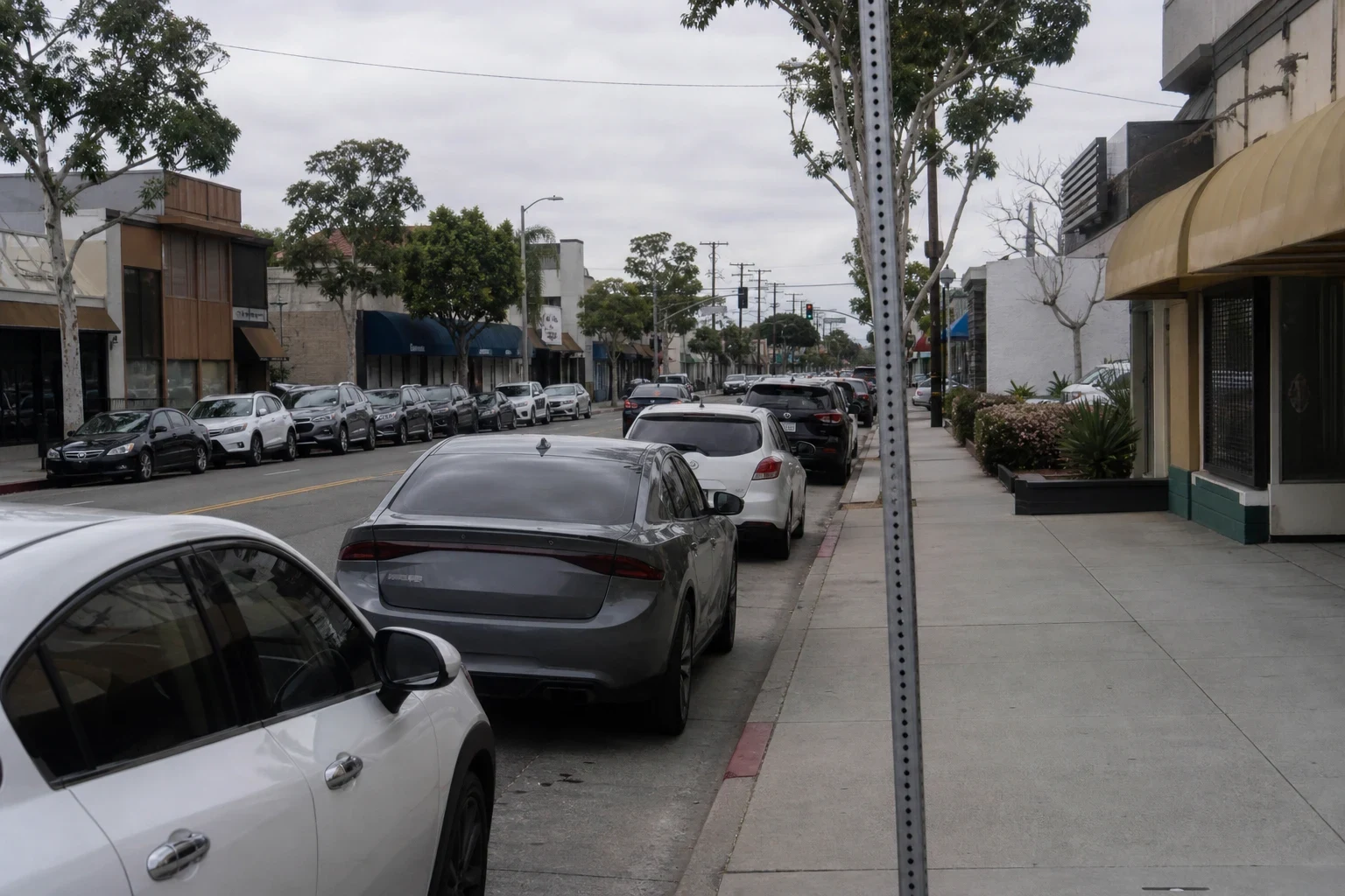 Overcast downtown street with low-rise shops, awnings and trees, cars parked along both curbs and a wide empty sidewalk on the right